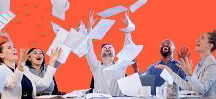 Group of joyful office workers tossing papers into the air, celebrating around a conference table, with a bright orange background.