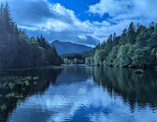 A scenic photo of Glencoe Lochan with tall trees and a distant mountain surrounding a lake
