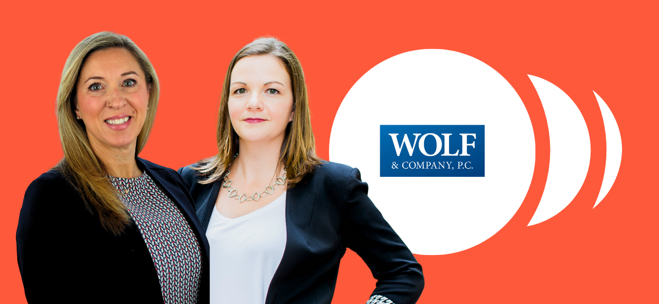 Two professional women standing in front of a bright orange background, next to the Wolf & Company, P.C. logo displayed inside a white circle.