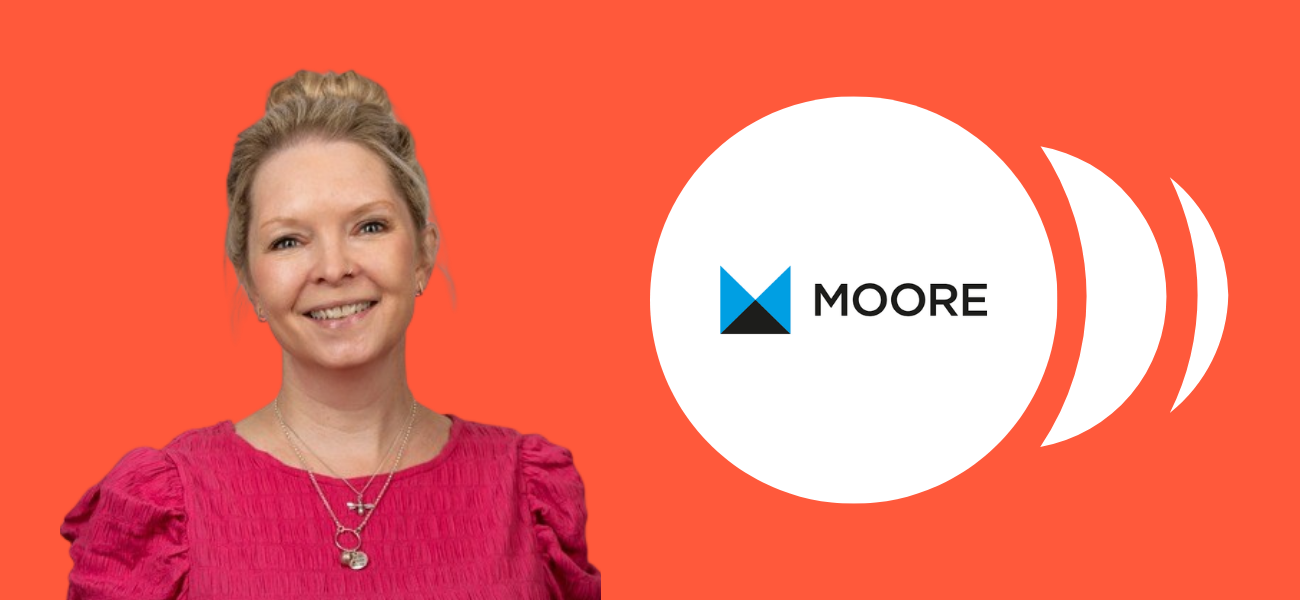 Smiling woman in a pink top standing in front of an orange background, next to the Moore logo displayed inside a white circle.