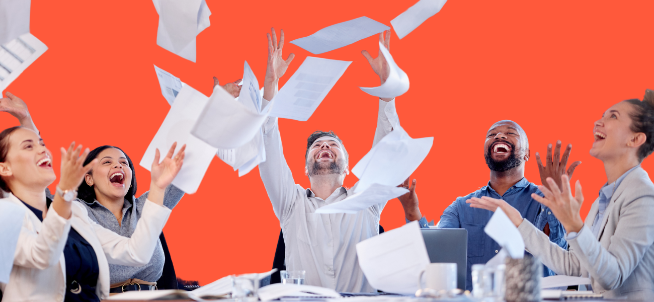 Group of joyful office workers tossing papers into the air, celebrating around a conference table, with a bright orange background.