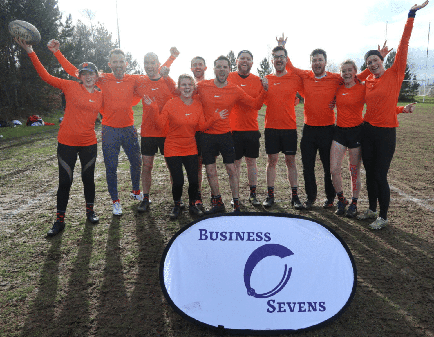 Dayshape touch rugby players in bright orange sports tops cheering in front of a Business Sevens sign